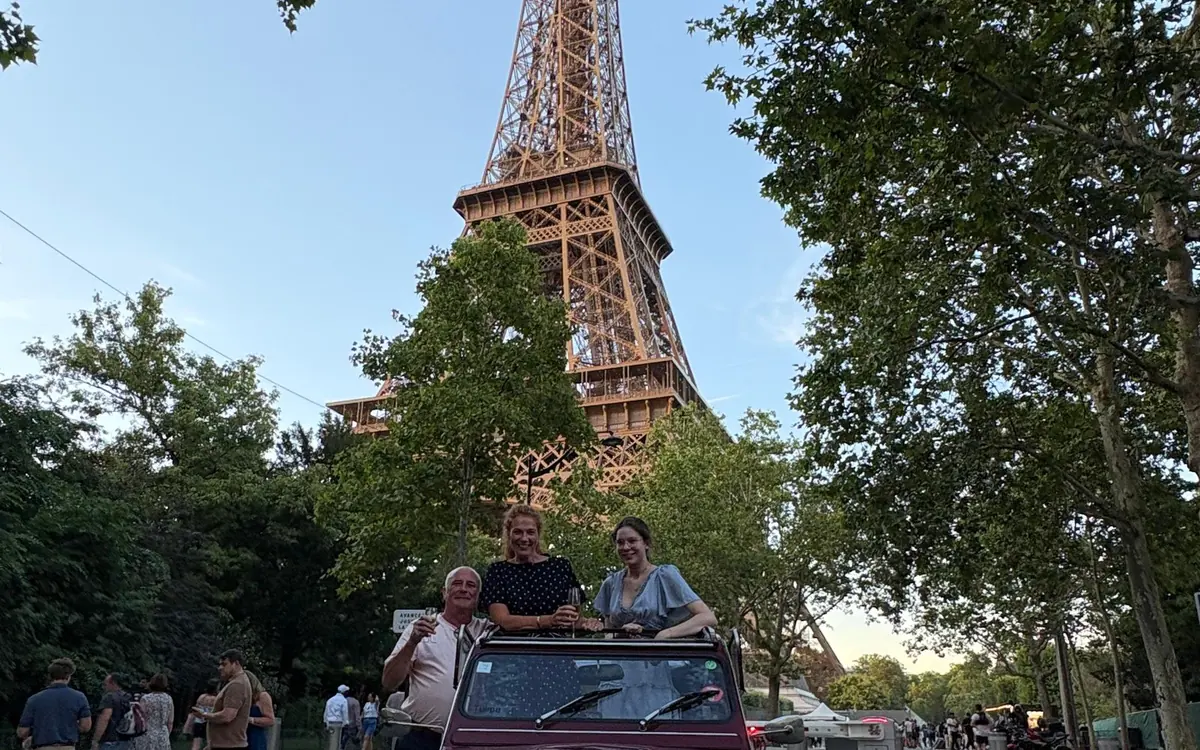 Three people in an open-top vintage Citroën 2CV parked under the Eiffel Tower