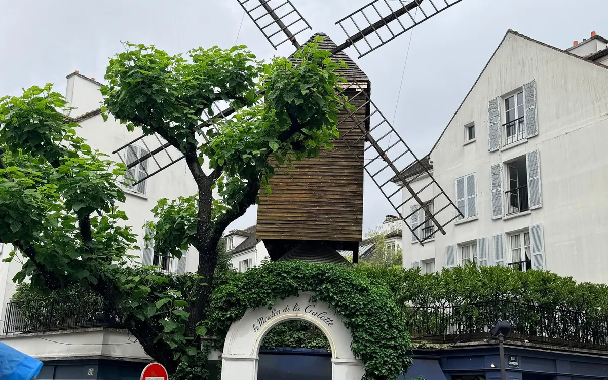 A wooden windmill behind leafy trees in a Montmartre street