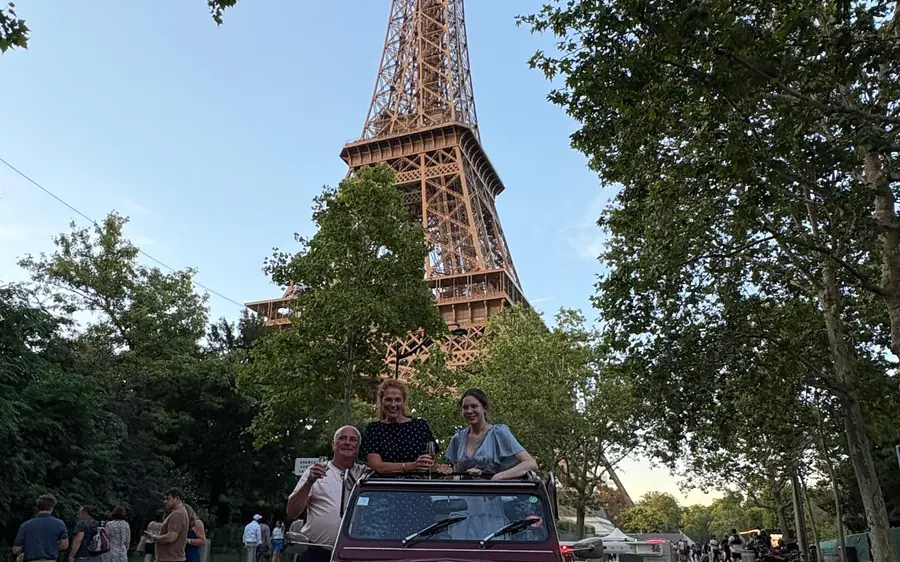 A vintage convertible with passengers parked below the Eiffel Tower